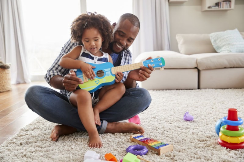 Photo of a father playing with his child with a toy guitar on the floor.