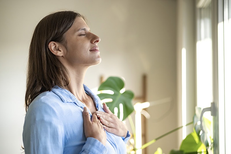 Woman taking a deep breath in her living room by the window in the sunlight.